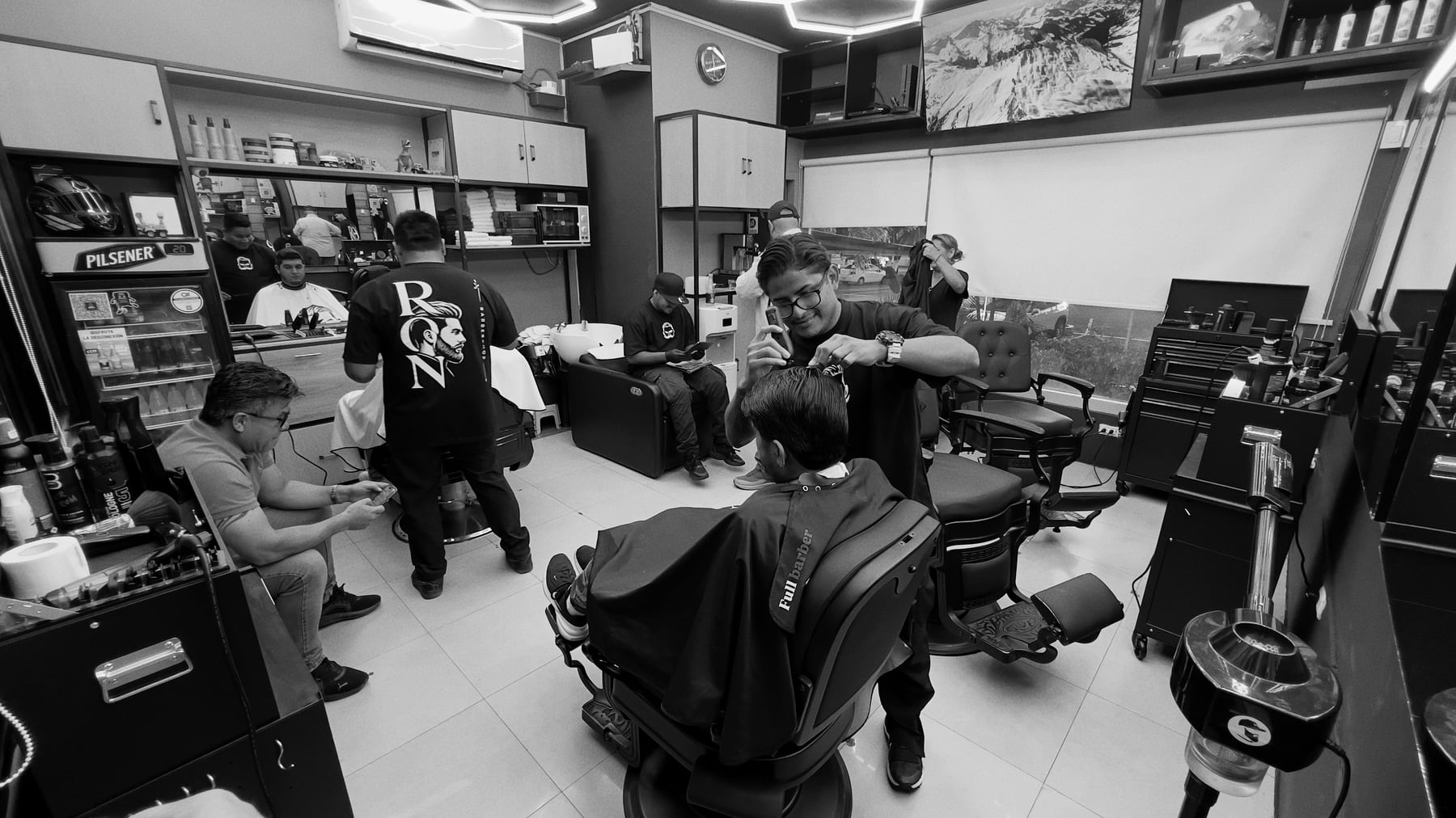 Clientes y barberos trabajando en las estaciones de Ron Barbershop.
